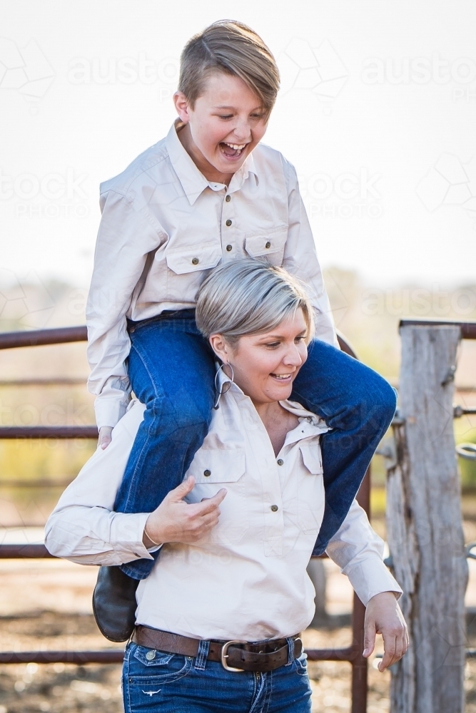 Single mum carrying son on shoulders laughing on farm in drought - Australian Stock Image