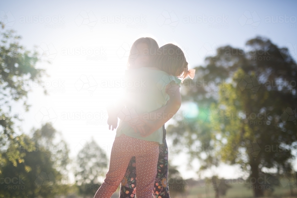 Sisters hugging with sunlight, trees and blue sky in the background - Australian Stock Image
