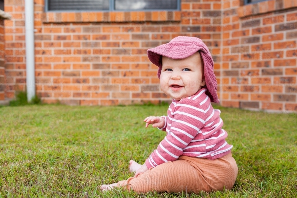 Six month old baby sitting on grass of front lawn beside home wearing hat - Australian Stock Image