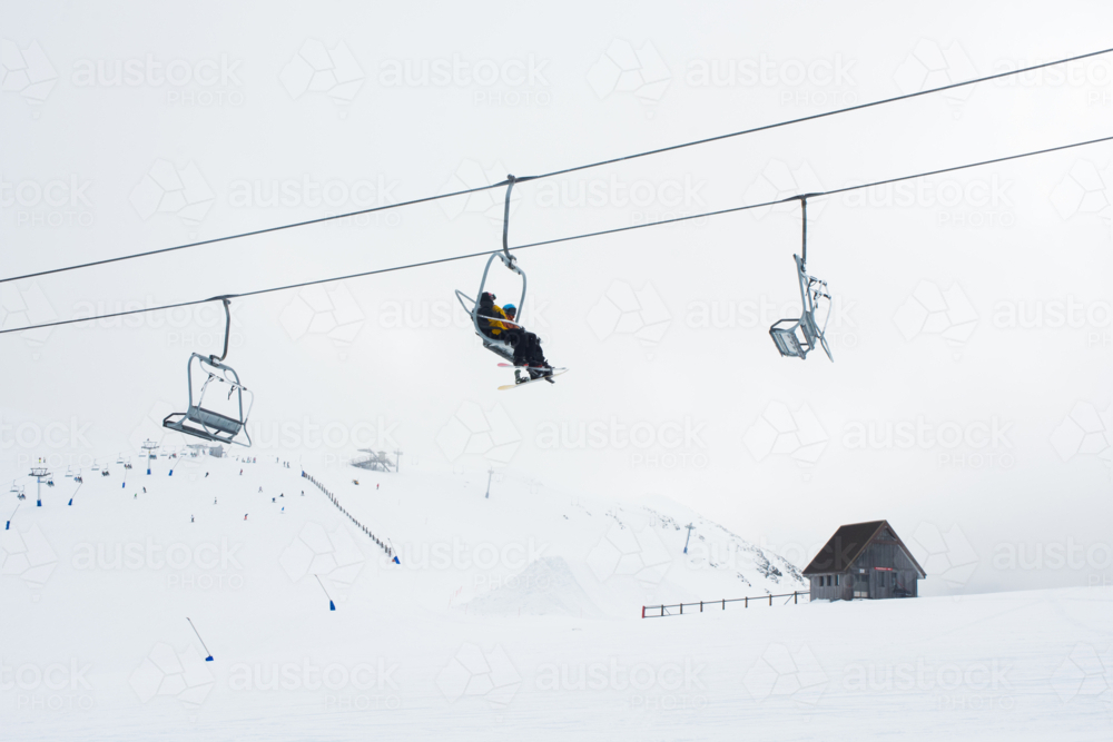 Skiers on ski lift with cabin and snowy mountain in background - Australian Stock Image