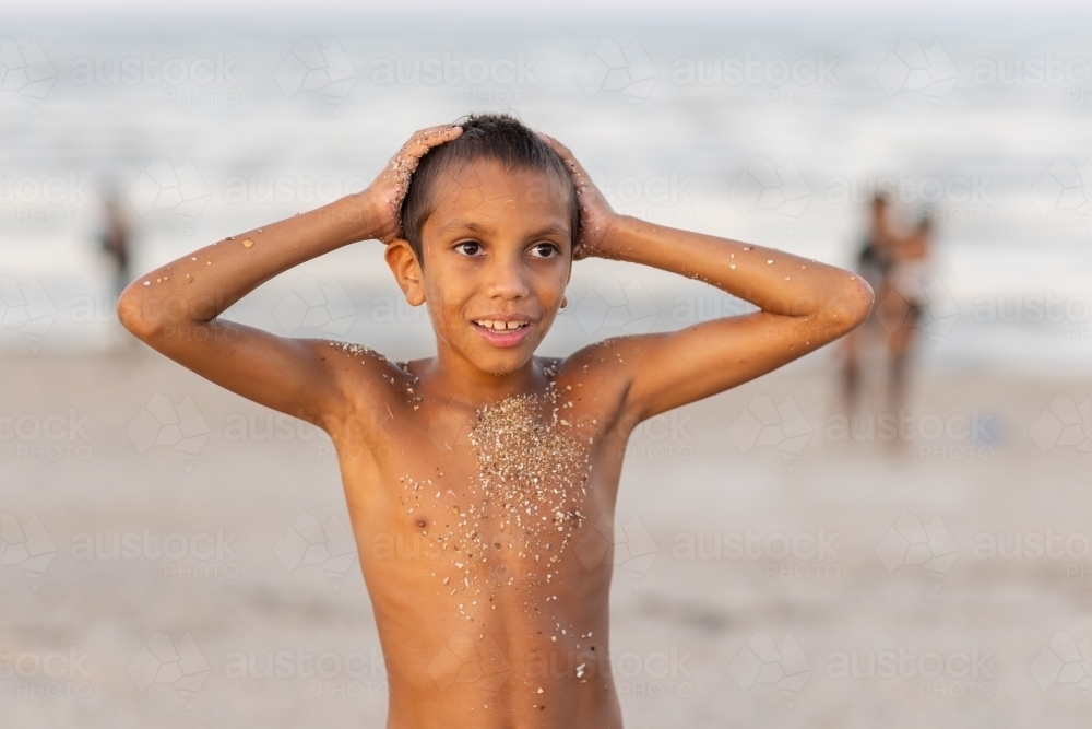 skinny aboriginal boy with hands on head covered in coarse sand on beach - Australian Stock Image