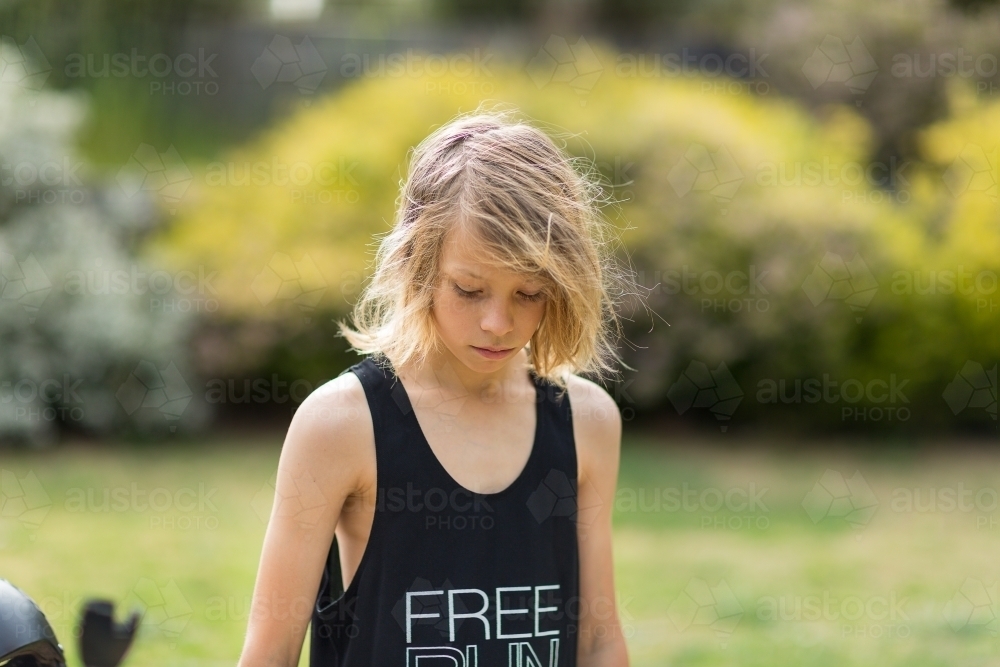 Skinny kid with scruffy hair and black singlet looking down - Australian Stock Image
