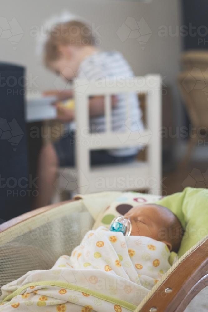 Sleeping infant wrapped in bassinet with older sibling in background - Australian Stock Image
