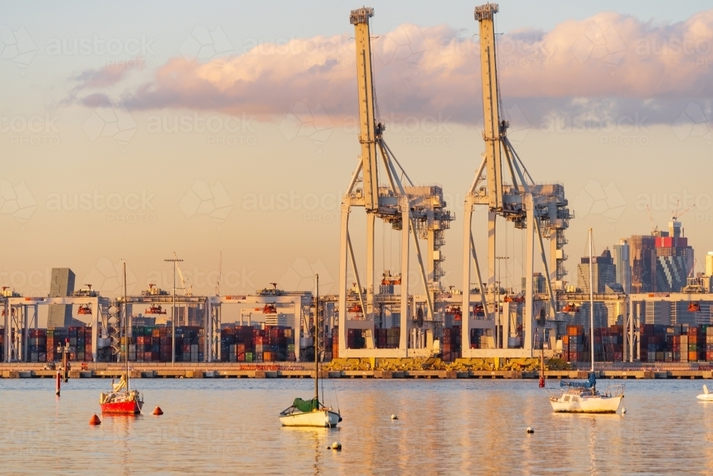 Small boats anchored in front of large gantry cranes at a city port at sunset. - Australian Stock Image