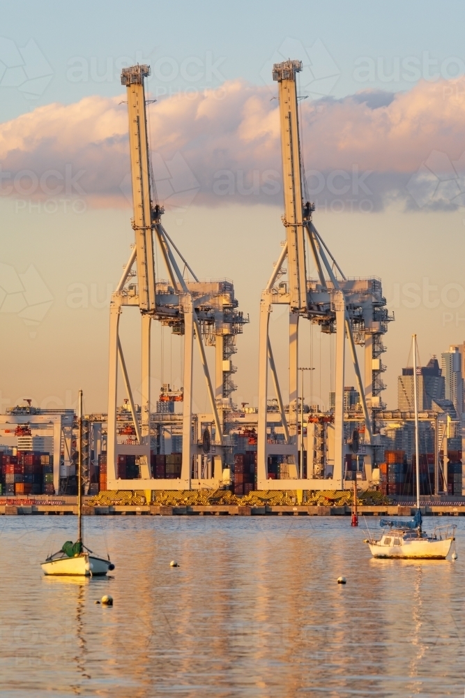 Small boats anchored in front of large gantry cranes at a city port at sunset. - Australian Stock Image
