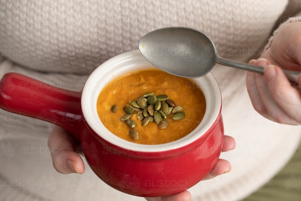 small red bowl of pumpkin soup in lady's hands with pepitas - Australian Stock Image