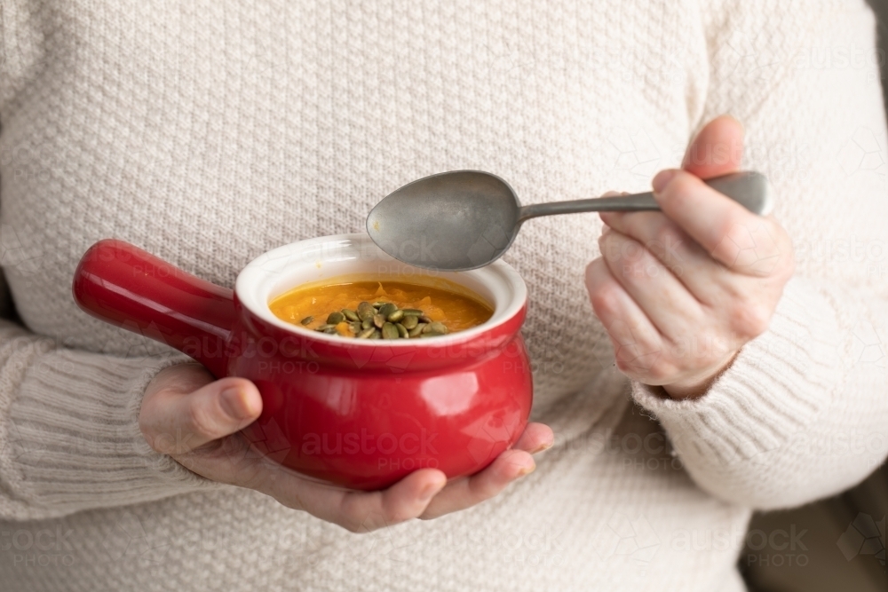 small red bowl of pumpkin soups in lady's hands with spoon - Australian Stock Image