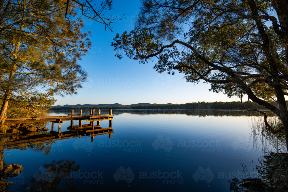Small timber jetty at sunrise beside calm water - Australian Stock Image