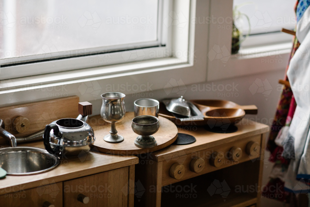 Small wooden play kitchen sits by a bright window with miniature cookware and utensils - Australian Stock Image