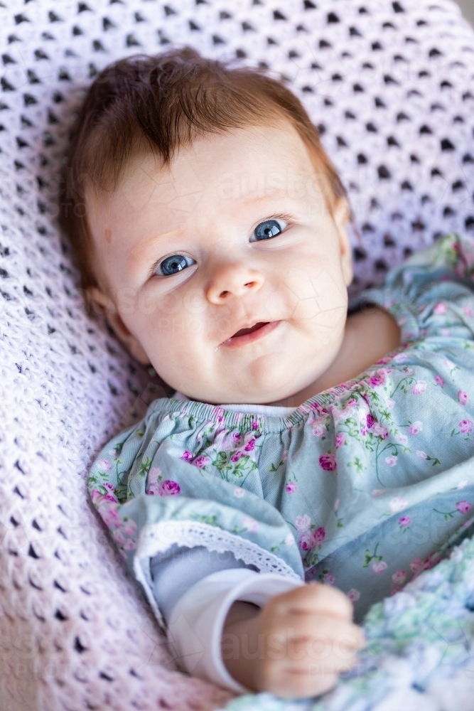smiling 9 week old baby with wide eyes sitting in bouncer on soft pastel pink crocheted blanket - Australian Stock Image