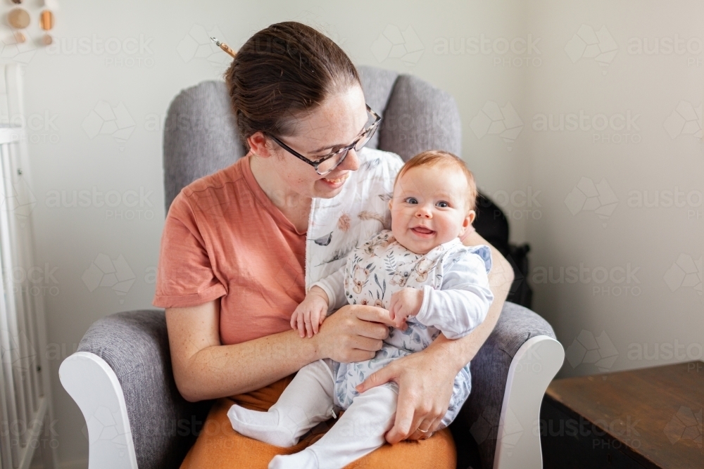 Smiling baby sitting on mums knee in rocking chair as they play together - Australian Stock Image