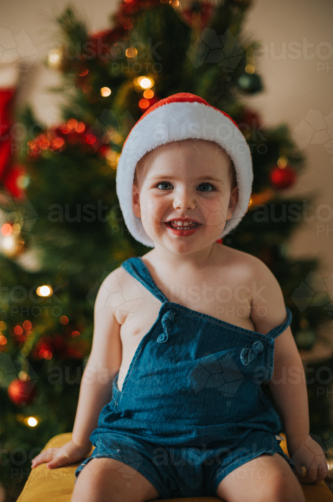 Smiling blonde girl in overalls sits cheerfully on a yellow stool in a Christmas setting - Australian Stock Image