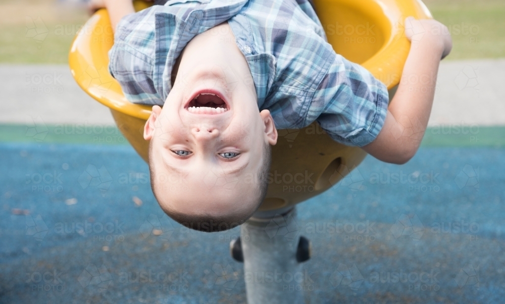 Smiling boy hanging upside down on play equipment at park. - Australian Stock Image