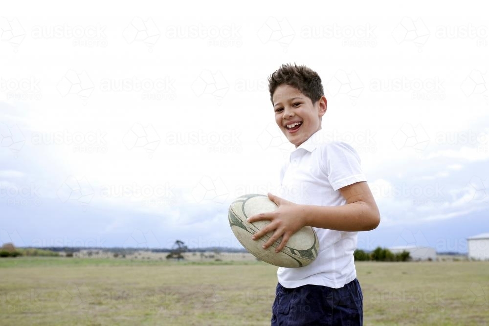 Smiling boy holding a rugby ball in a paddock - Australian Stock Image