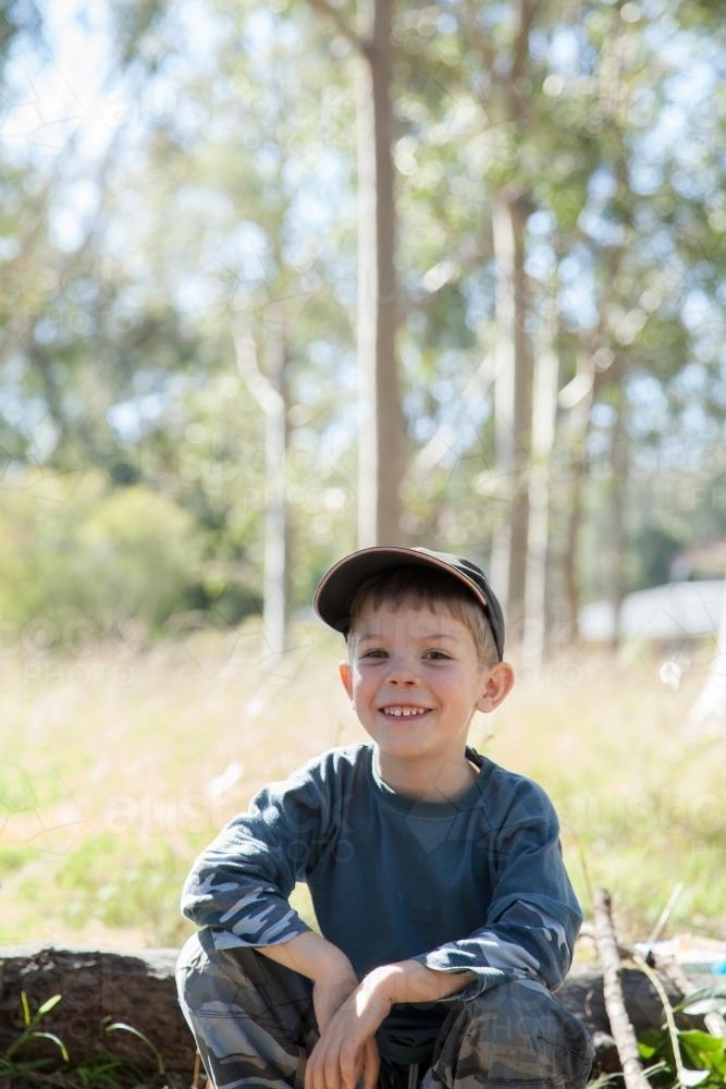 Smiling boy sitting on a log in a paddock by a campfire - Australian Stock Image