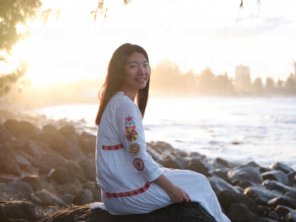 Smiling Chinese woman sitting on rocks by the shore in the evening - Australian Stock Image