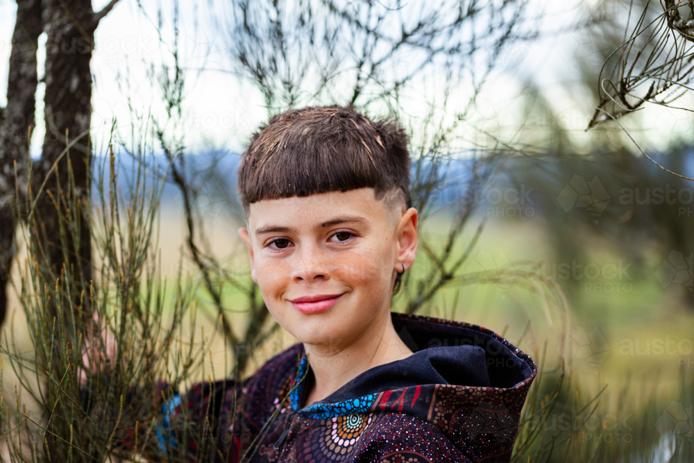 Smiling close up portrait of aboriginal boy wearing ochre by casuarina tree - Australian Stock Image