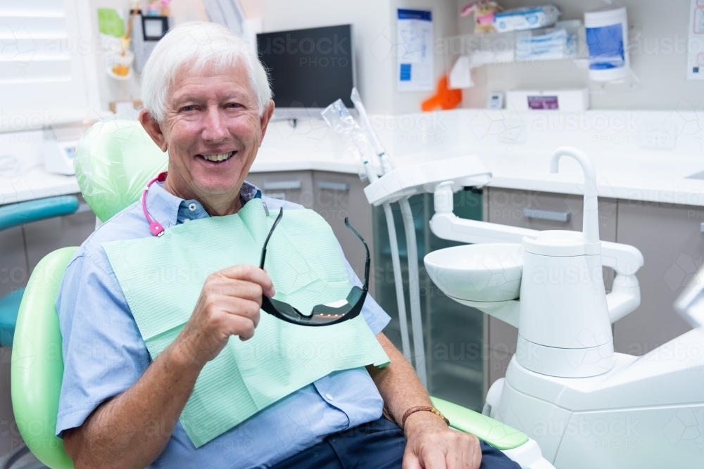 Smiling elderly male patient in dental office holding safety glasses - Australian Stock Image