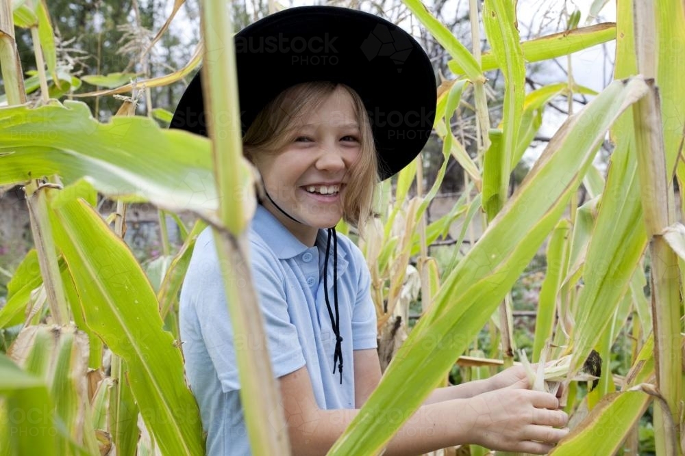 Smiling girl wearing school uniform and hat standing in the veggie patch - Australian Stock Image