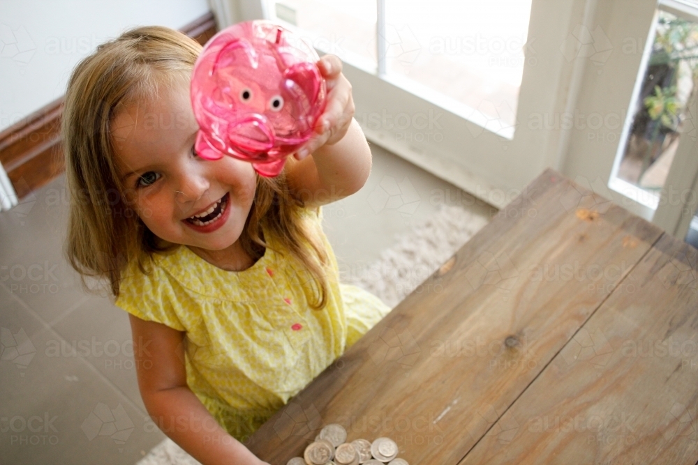 Smiling girl with blonde hair wearing yellow blouse raising a pink transparent piggy bank - Australian Stock Image