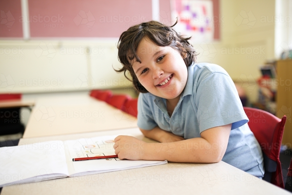 smiling girl with short hair wearing pale blue shirt sitting with a book and pencil on the table - Australian Stock Image