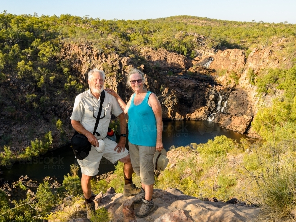 Smiling grey haired man and woman posing on rock with scenic waterfall and plunge pool behind - Australian Stock Image
