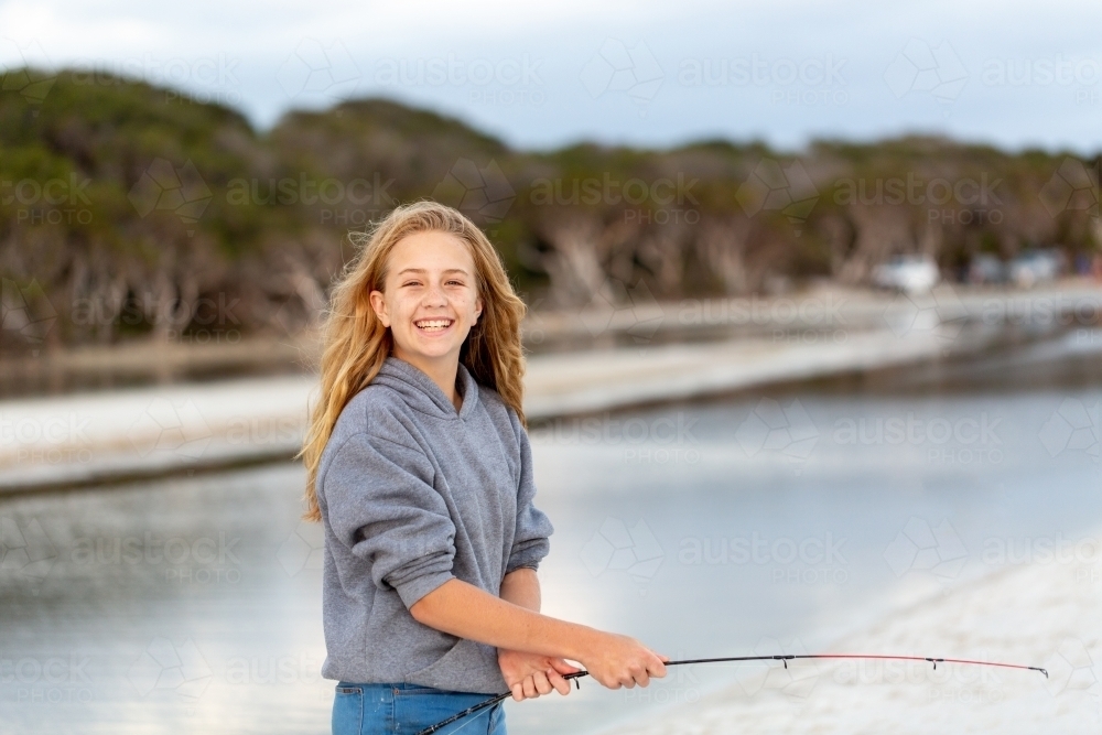 Smiling happy girl on beach in front of water with fishing rod - Australian Stock Image
