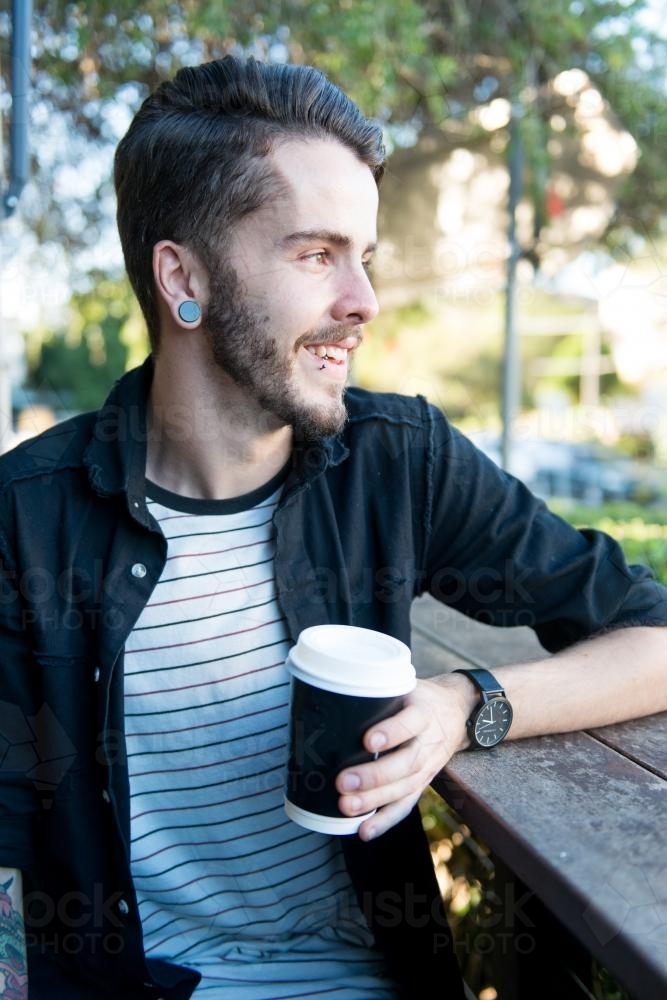 Smiling hipster man sitting outdoors with a takeaway coffee - Australian Stock Image