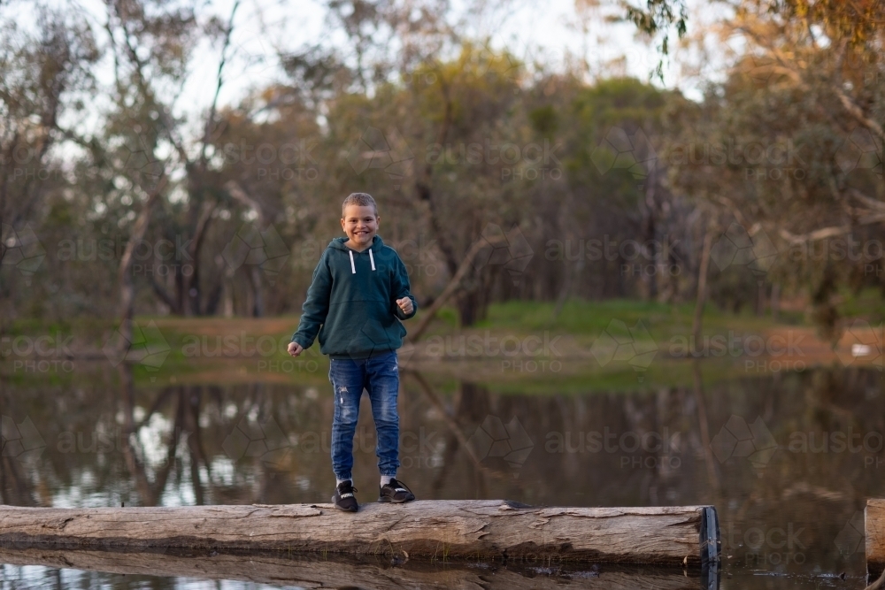 smiling kid standing on log at edge of lake - Australian Stock Image