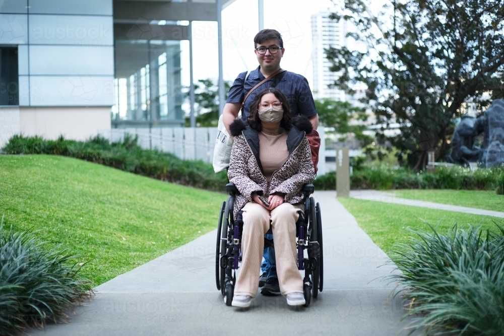Smiling man standing behind the woman with a disability with face mask outdoors - Australian Stock Image