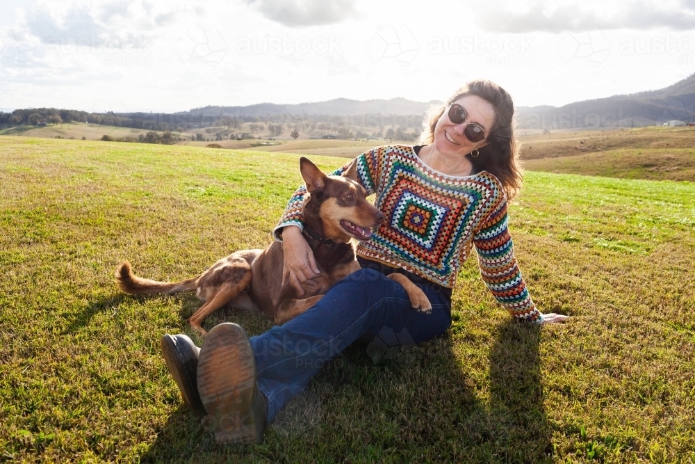 Smiling middle age Aussie woman sitting on farm lawn with pet kelpie dog - Australian Stock Image
