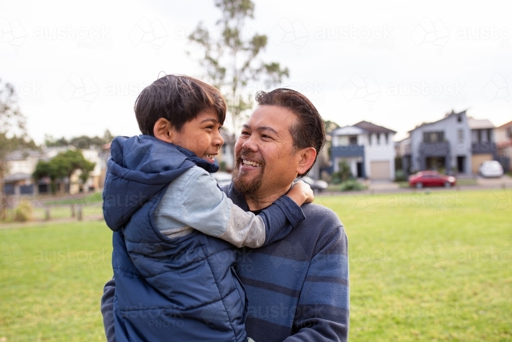 smiling middle aged man wearing blue sweatshirt carrying a smiling boy in his arms on a big lawn - Australian Stock Image