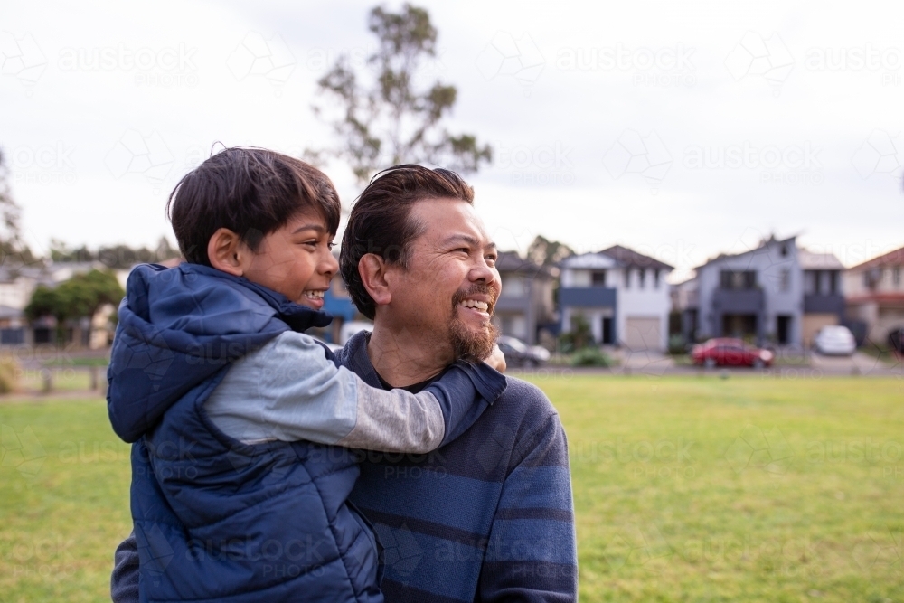 smiling middle aged man wearing blue sweatshirt carrying a smiling boy in his arms on a big lawn - Australian Stock Image