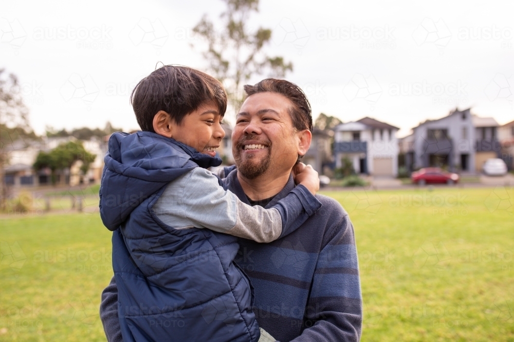 smiling middle aged man wearing blue sweatshirt carrying a smiling boy in his arms on a big lawn - Australian Stock Image