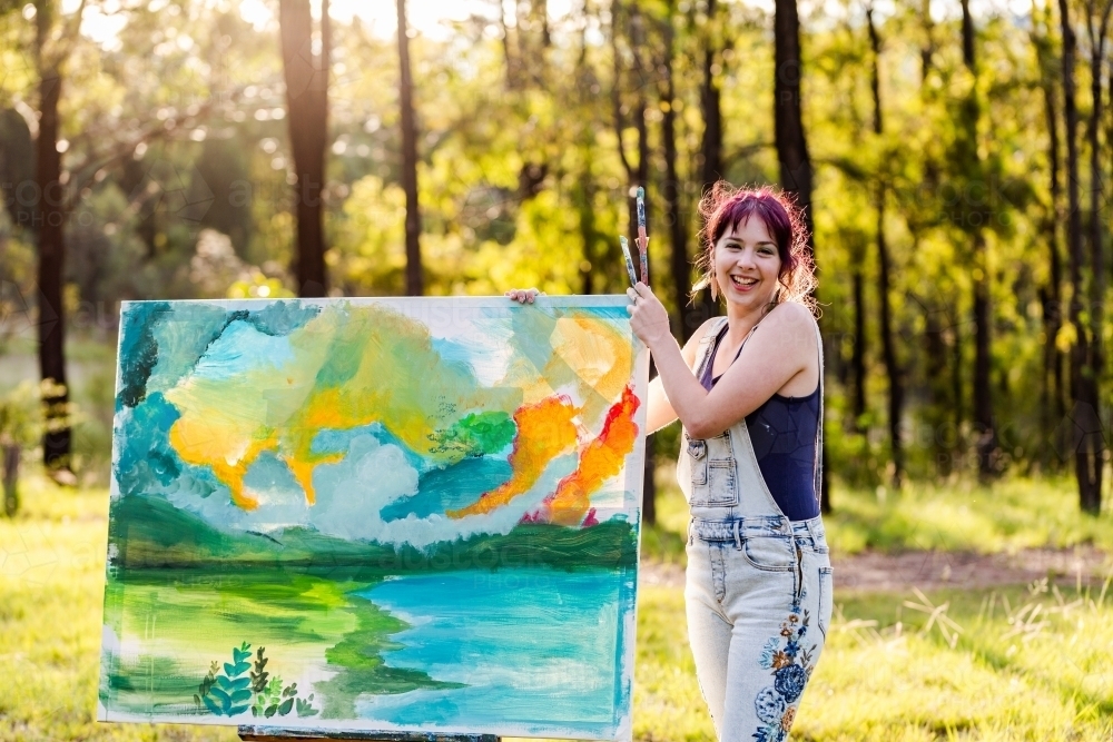 Smiling portrait of happy young woman artist in forest clearing with beginning of painting - Australian Stock Image