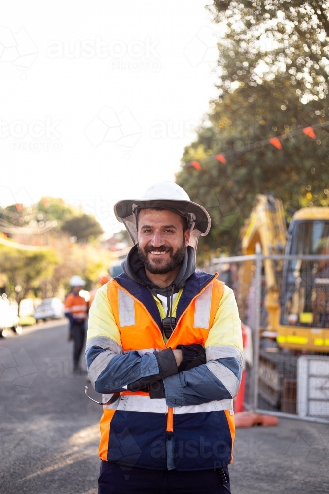Smiling road worker man with beard wearing orange and yellow high-vis jacket with his arms crossed - Australian Stock Image