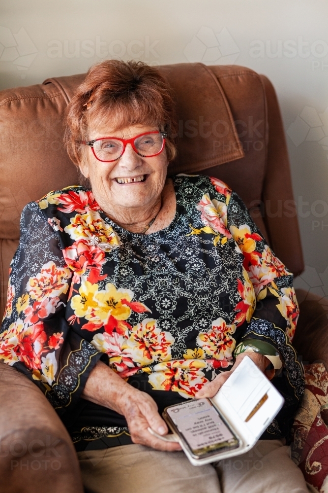 Smiling senior woman sitting in chair using mobile phone wearing glasses - Australian Stock Image