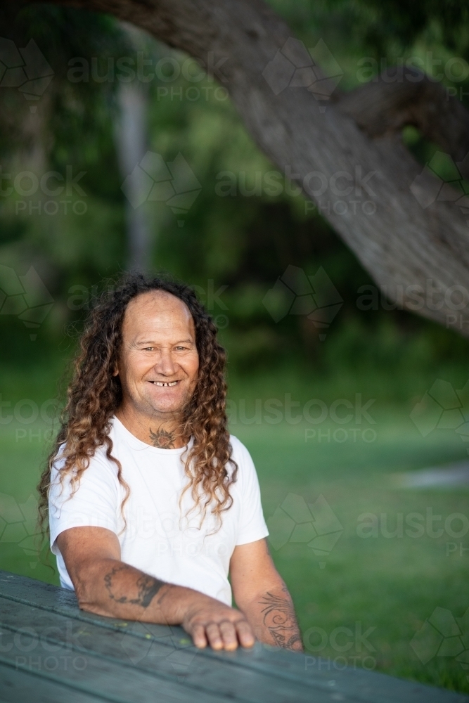 smiling tattooed man in white tee-shirt with long curly hair - Australian Stock Image