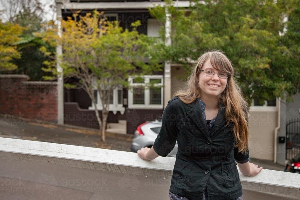 Smiling teenage girl with glasses leaning back against railing along the street - Australian Stock Image