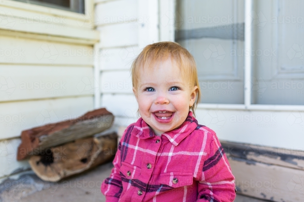 Smiling toddler girl portrait sitting on back doorstep of country farm house beside firewood - Australian Stock Image