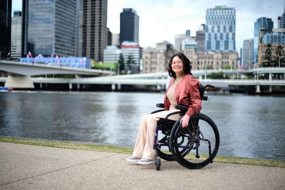 Smiling woman in wheelchair by river with city in background - Australian Stock Image