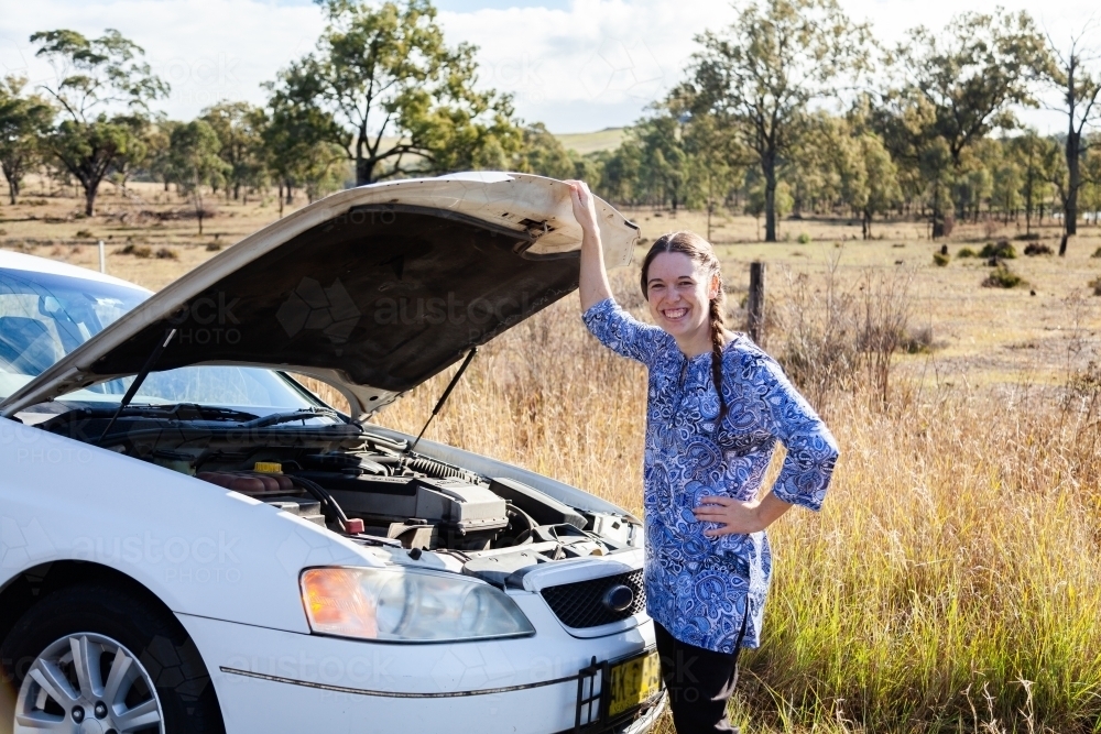 Smiling woman standing beside car stopped on roadside with bonnet open - Australian Stock Image