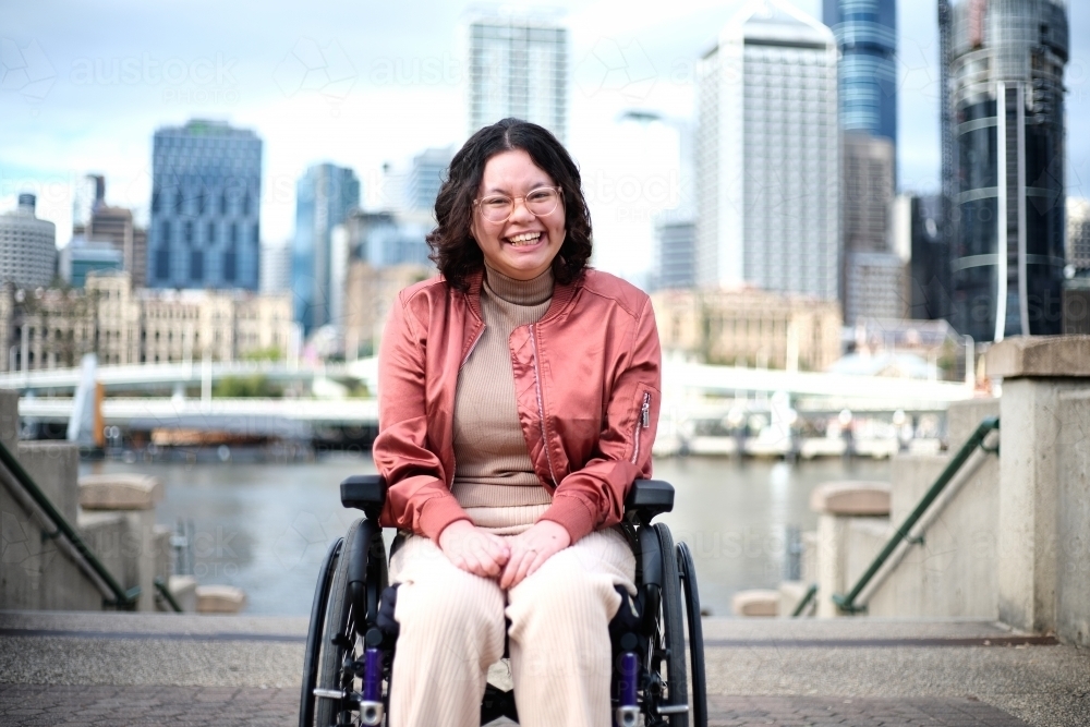 Smiling woman with a disability sitting in a wheel chair with tall buildings behind her - Australian Stock Image