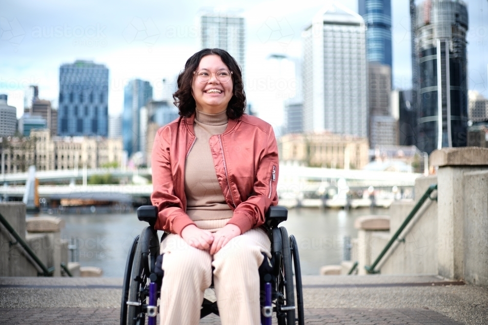 Smiling woman with a disability sitting in a wheel chair with tall buildings behind her - Australian Stock Image