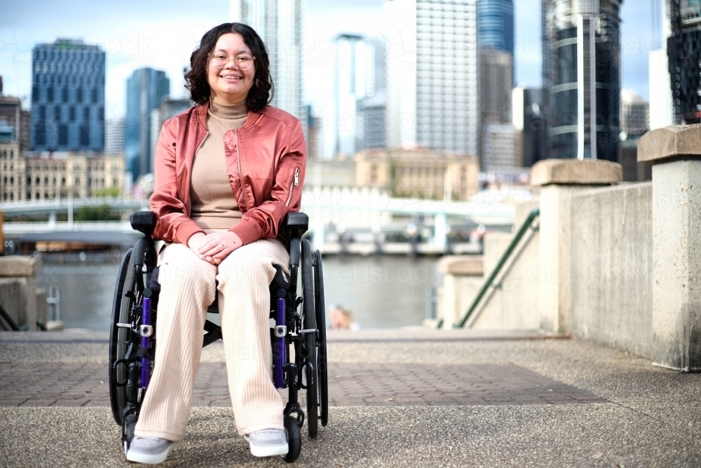 Smiling woman with a disability sitting in a wheel chair with tall buildings behind her - Australian Stock Image