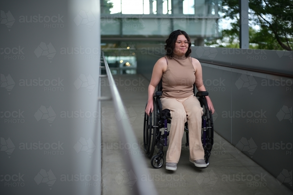 Smiling woman with a disability sitting in a wheelchair going down an accessible ramp - Australian Stock Image