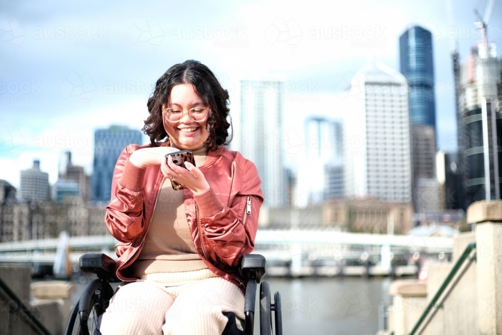 Smiling woman with a disability sitting in a wheelchair in the city with mobile phone - Australian Stock Image