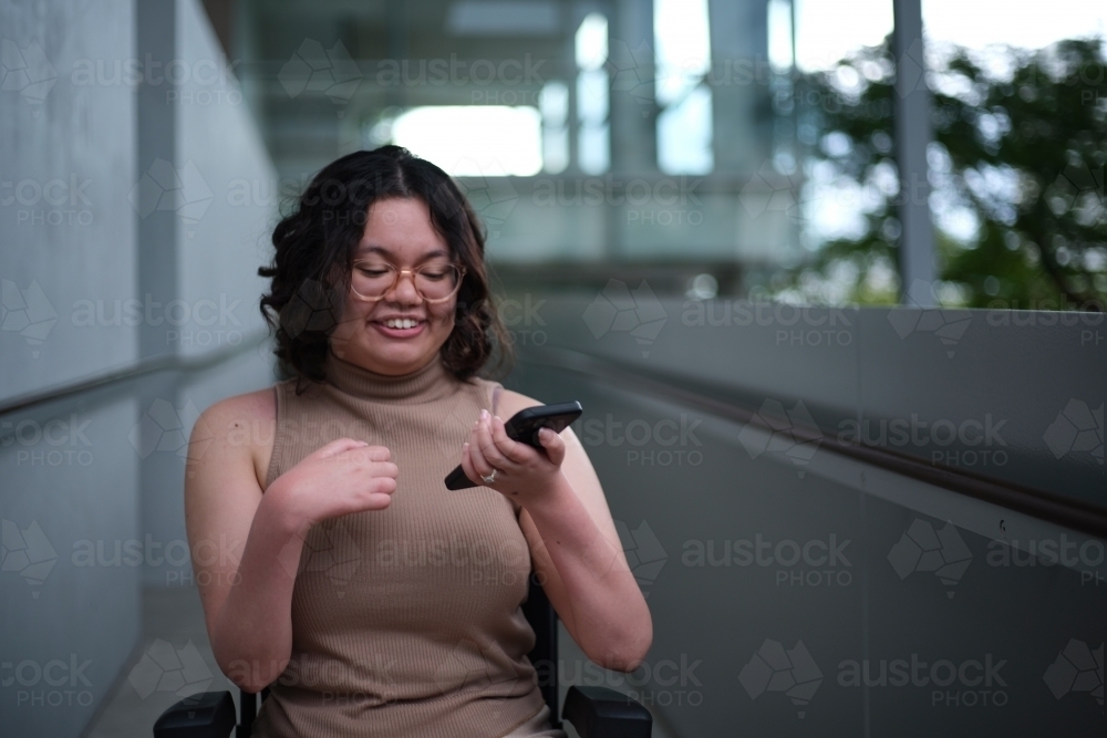 Smiling woman with a disability sitting in a wheelchair indoors holding a mobile phone - Australian Stock Image