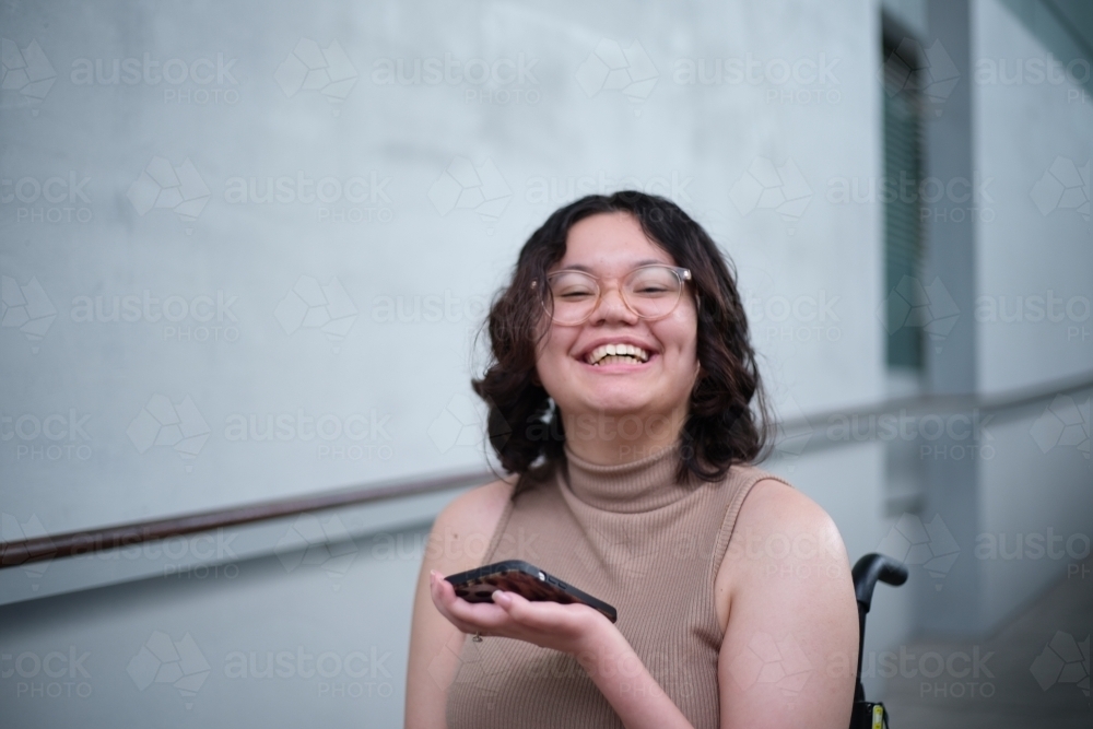 Smiling woman with a disability sitting in a wheelchair indoors holding a mobile phone - Australian Stock Image