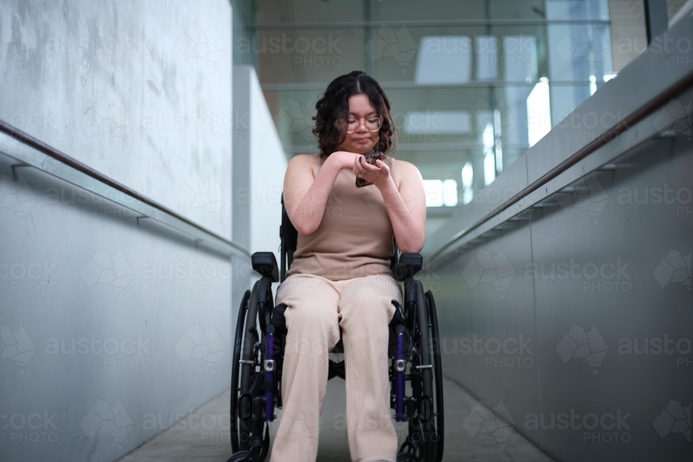 Smiling woman with a disability sitting in a wheelchair looking down at her mobile phone - Australian Stock Image